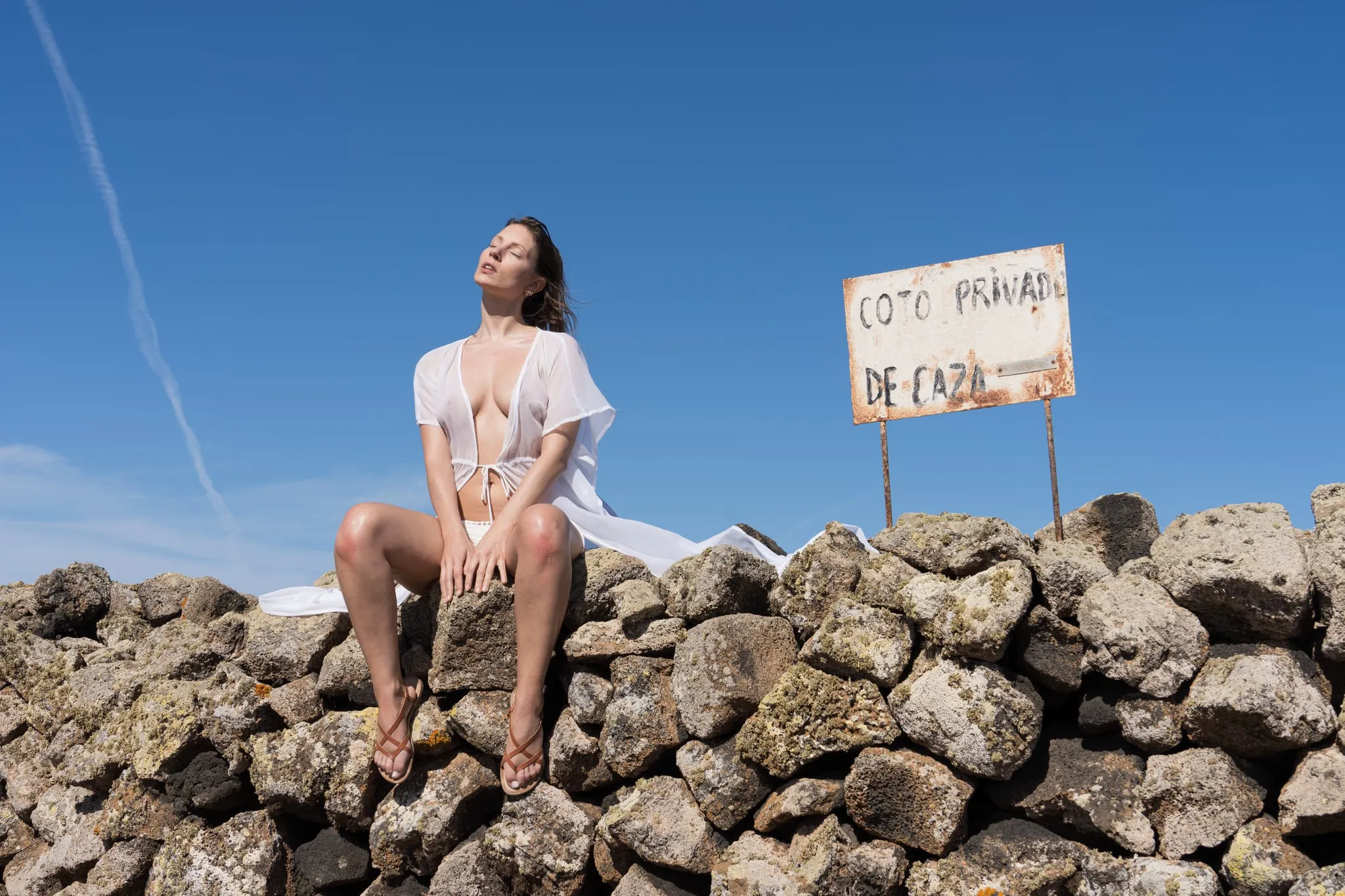 Model in sheer white sitting on a volcanic stone wall under deep blue sky, a weathered Spanish sign behind