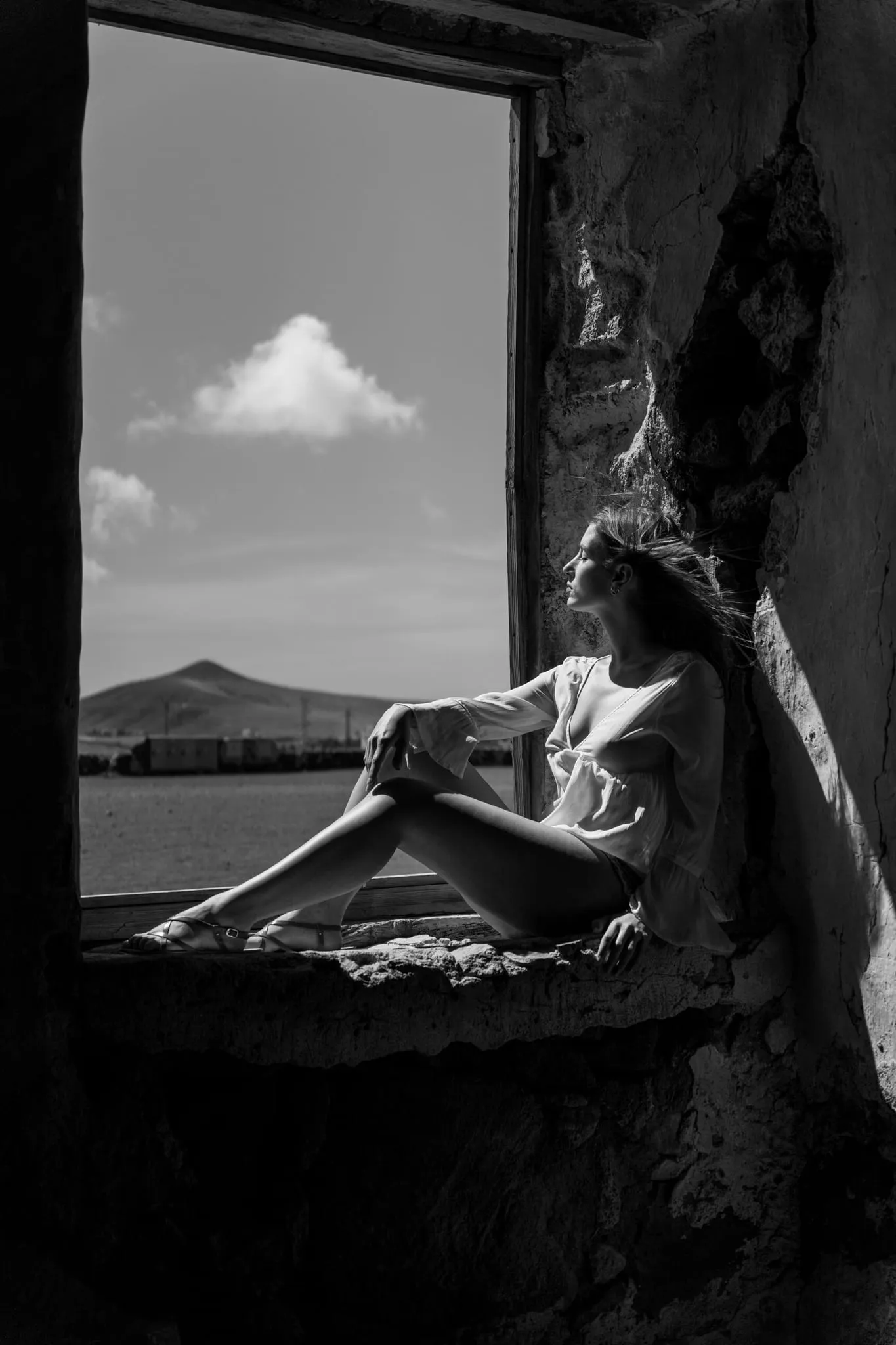 Black-and-white portrait of a woman seated in a ruined stone window, a volcanic hill framed beyond