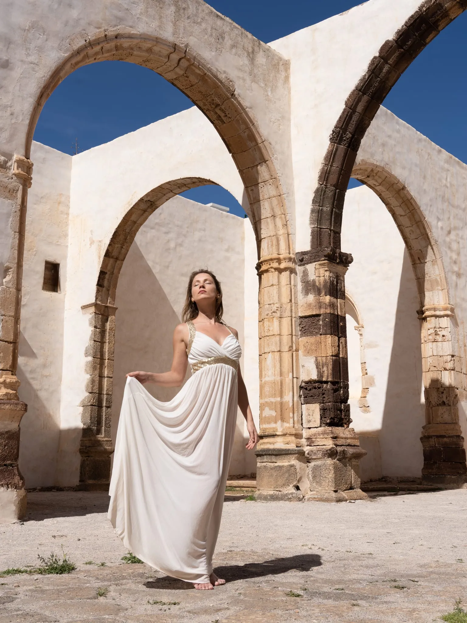 Woman in a flowing white dress walking barefoot among sunlit stone arches of an old convent
