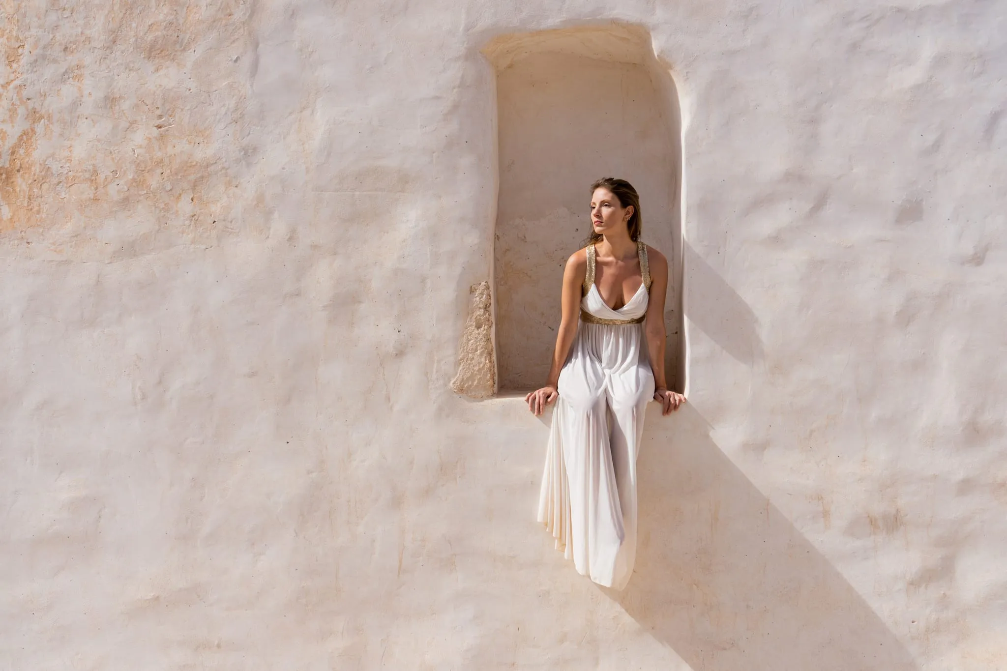 Woman in a white gown seated in the niche of a sunlit whitewashed wall