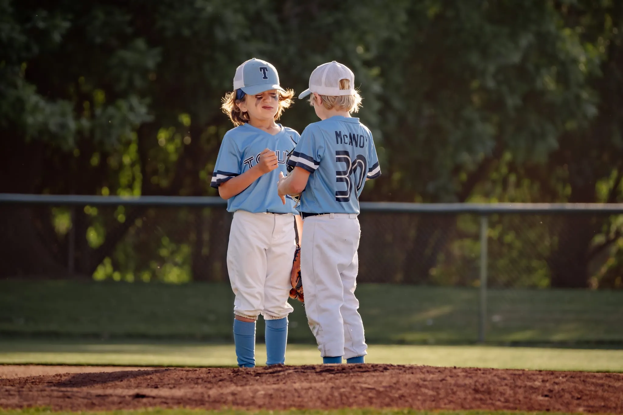 Two young teammates confer on the pitcher's mound in late-afternoon light