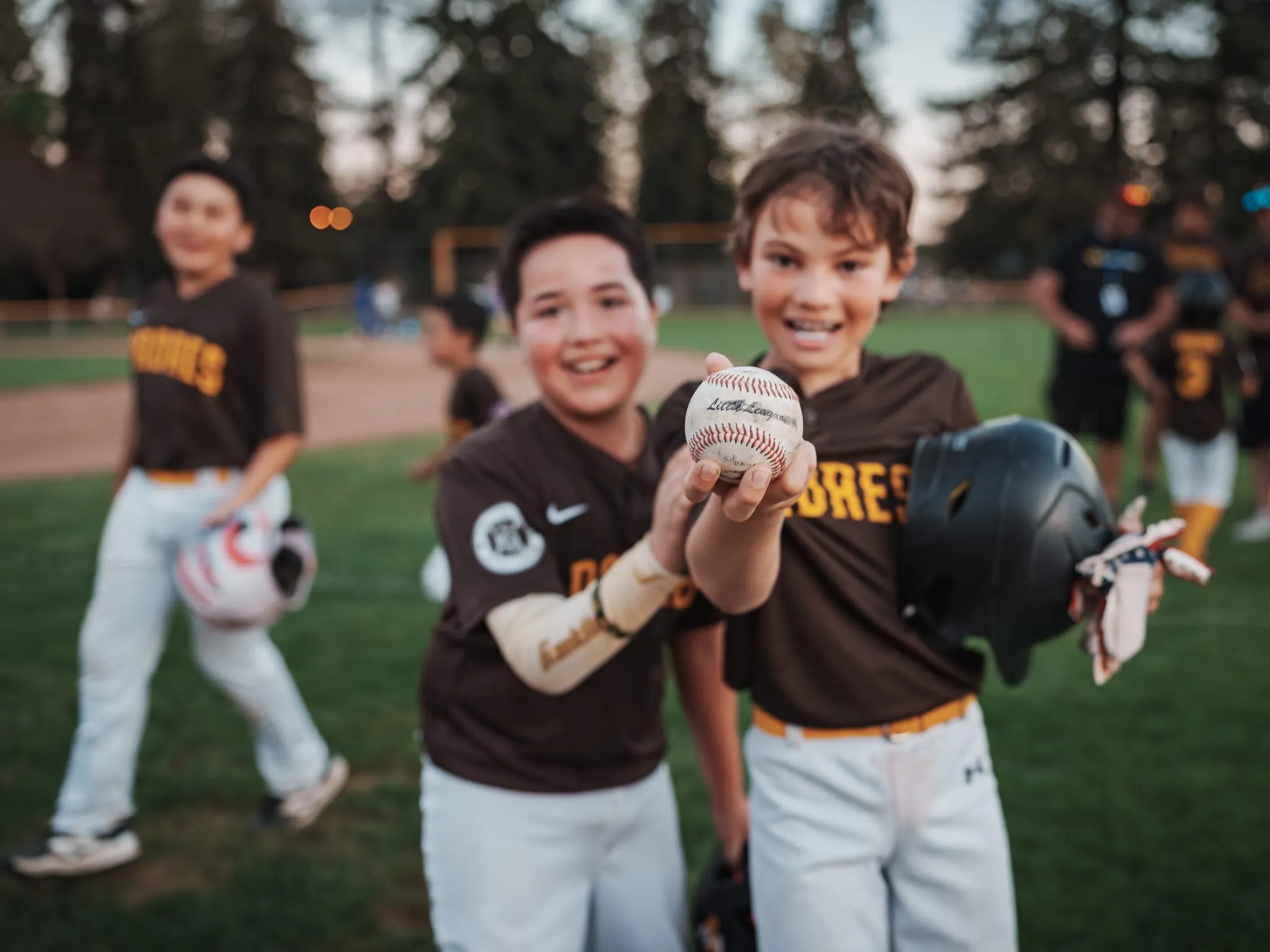 Two Little League players grin and hold up the game ball after an evening game on a Bay Area field