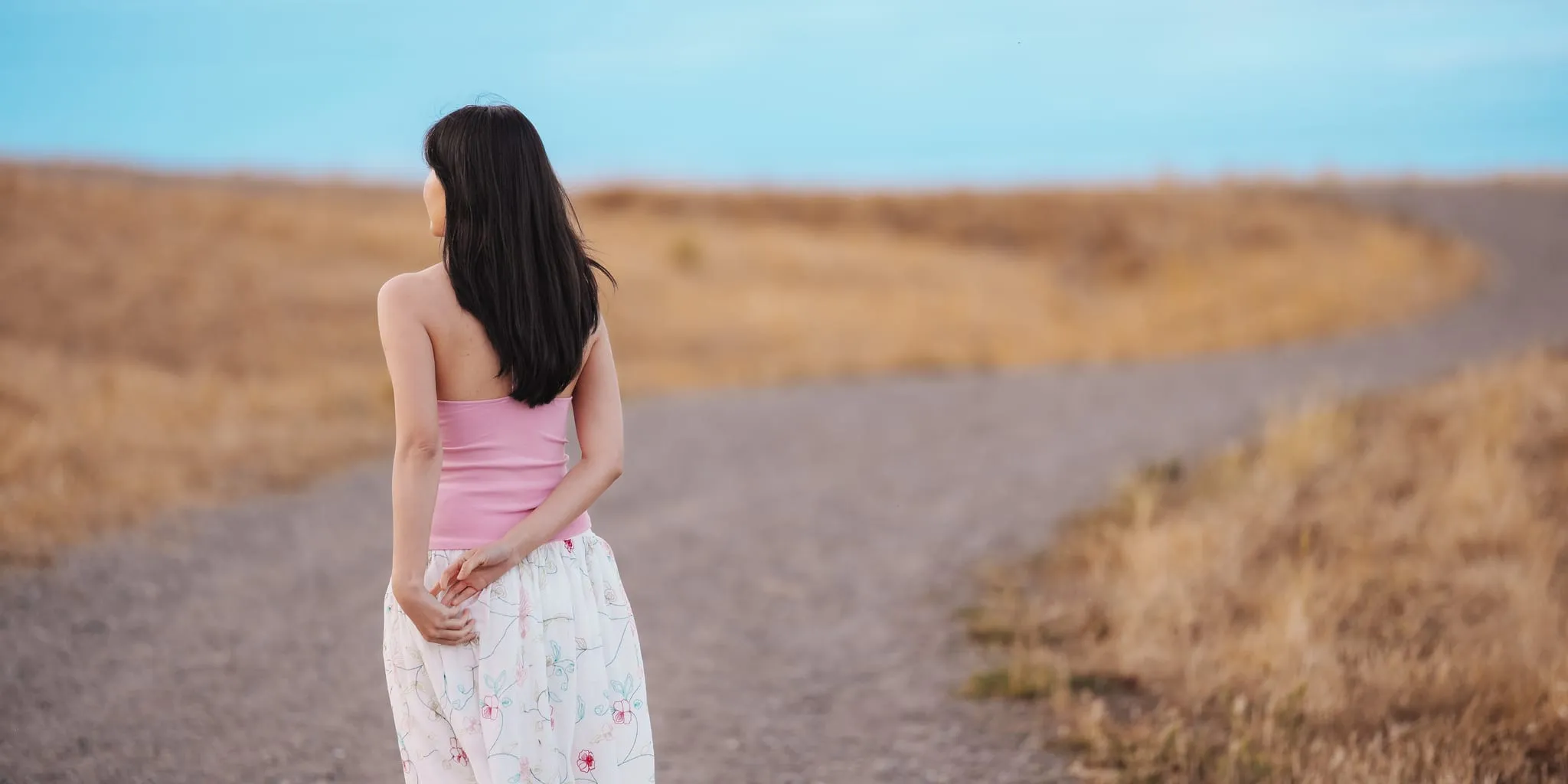 Seen from behind, a woman pauses where a gravel path curves through golden grassland