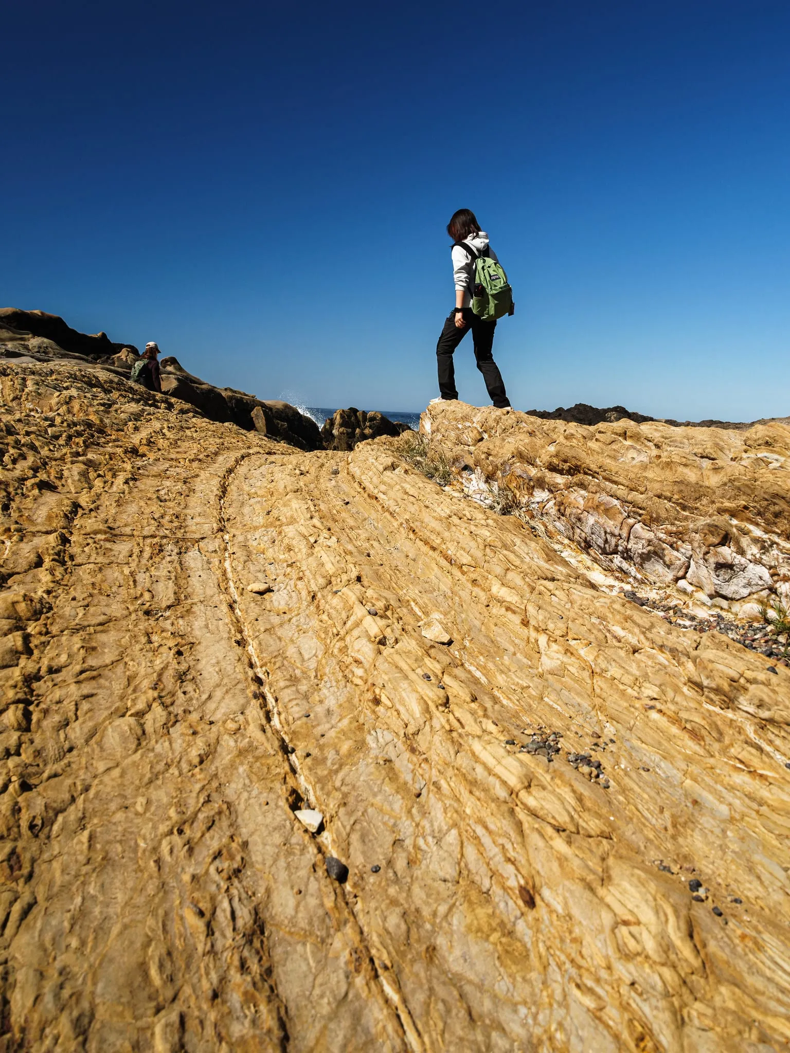 A hiker stands on striated golden sandstone above the surf under a deep blue sky on the California coast